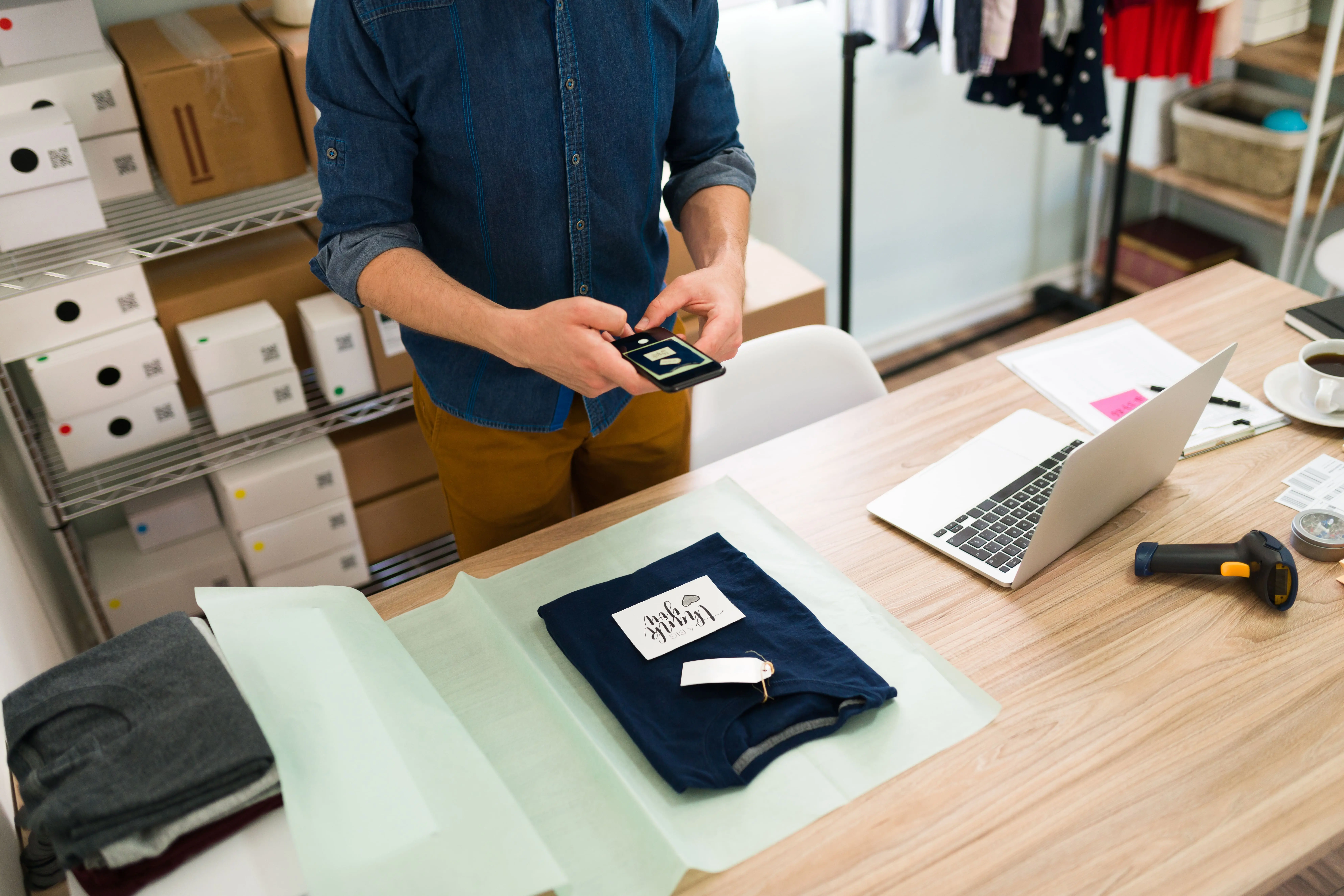 A person in a denim shirt is scanning a clothes tag with their phone at a desk with folded clothes, a laptop, and a barcode scanner, surrounded by shelves of boxes and hanging clothes. They're checking the MCC to determine the Merchant Category Codes.